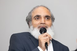A person with a white beard holding a microphone

Description automatically generated